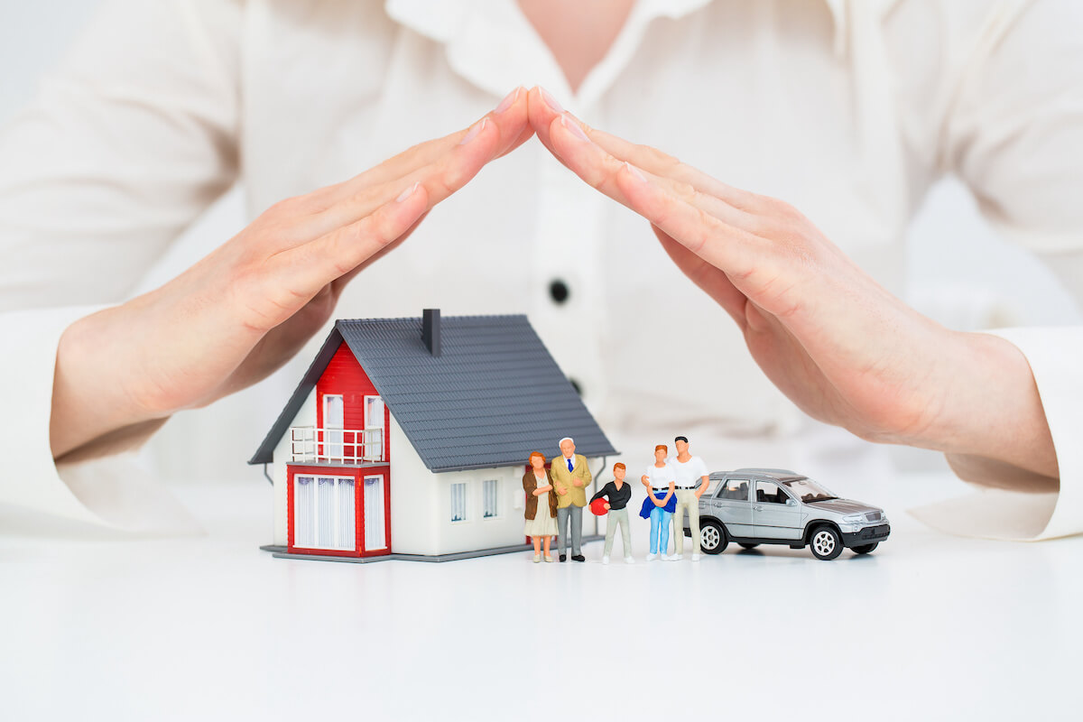 hands covering a model house, car, and family like a shield